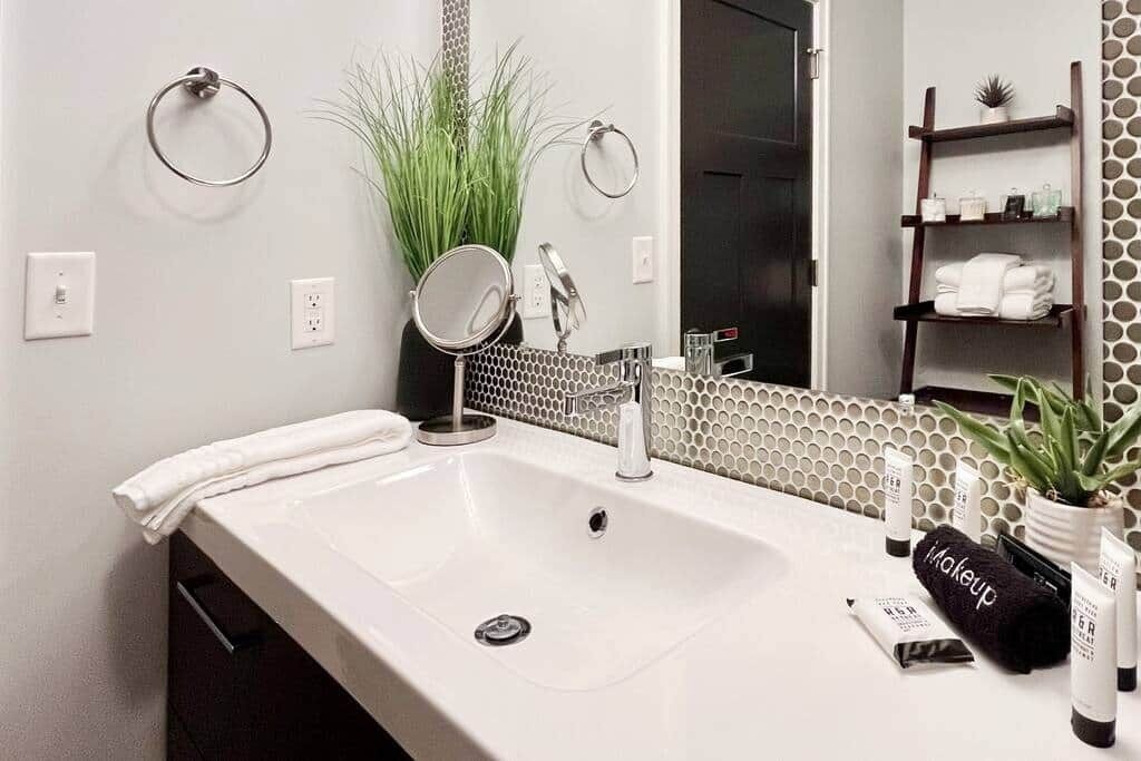 A modern bathroom with a white sink, a round mirror, a towel ring, a potted plant, and a shelf with toiletries.
