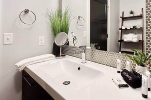 A modern bathroom with a white sink, a round mirror, a towel ring, a potted plant, and a shelf with toiletries. - Sunsets on the Grand (Lansing)