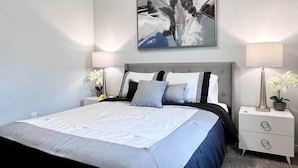 A modern bedroom with a large bed, two bedside tables with lamps, a wall-mounted artwork, and a potted plant. - Sunsets on the Grand (Lansing)