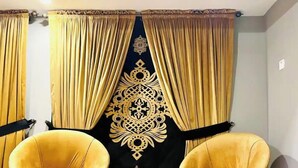 Ein Zimmer mit zwei gelben Sesseln und einem passenden Hocker, einer dekorativen, schwarzen Wandteppich mit Goldmustern und goldenen Vorhängen.