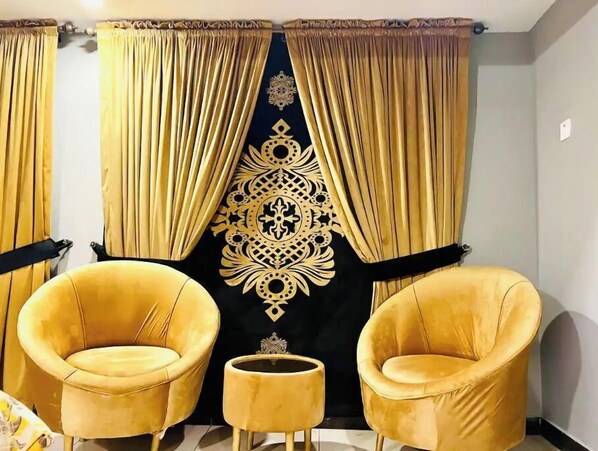 A room with two yellow armchairs and a matching ottoman, a decorative black tapestry with gold patterns, and golden curtains.