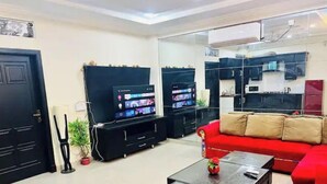 Un salon avec un canapé rouge, deux meubles de rangement noirs pour appareils électroniques, une table basse en verre et un coin cuisine équipé de placards et d’appareils électroménagers.