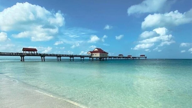 En strand med kristallklart turkost vatten, en sandstrand och en brygga med bungalows som är byggda på pålar över vattnet.