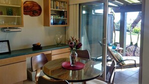 A dining area with a glass table, chairs, a vase with flowers, a ceiling fan, and a view of the outdoors through a sliding glass door. - #17 Colony One at Sea Mt. Punalu`u (Pahala)