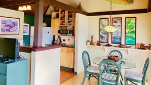 A kitchen with wooden cabinets, a microwave, and a dining area with a glass table and blue chairs. - #26 Colony One at Sea Mt. Punalu`u (Pahala)