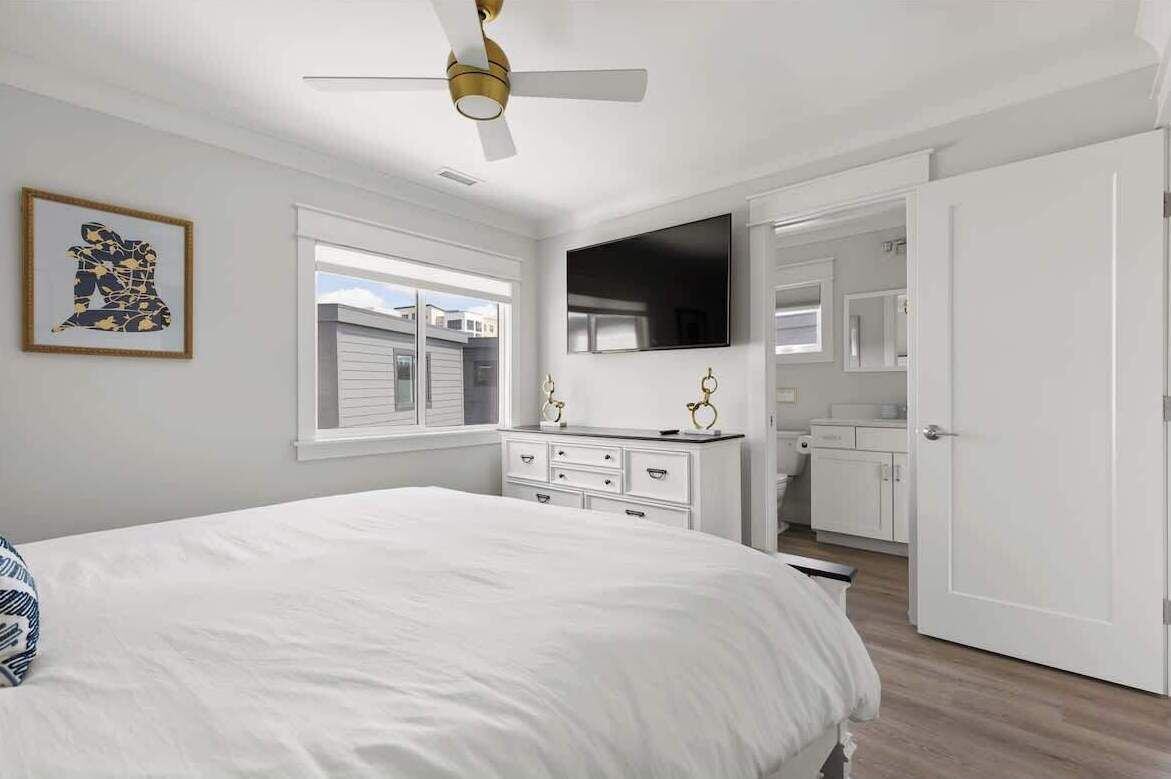 Une chambre à coucher comprenant un lit, un ventilateur de plafond, une télévision, une commode et une porte donnant sur une salle de bain.