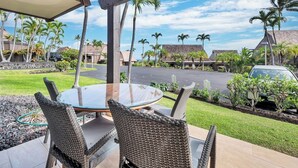 Un patio techado al aire libre con mobiliario de mimbre, una mesa de vidrio y vistas a una calle bordeada de palmeras y casas.