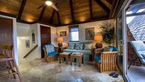 Un salon chaleureux avec un plafond haut, des poutres en bois, un ventilateur de plafond, un canapé vert agrémenté de coussins à motifs, une table basse et un fauteuil en osier.