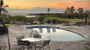 Un espace piscine avec des chaises longues et une table, offrant une vue sur l’océan et un magnifique coucher de soleil.