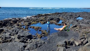 Ein felsiger Strand mit klarem, blauem Wasser, ein Kind steht dort, und eine Person sitzt auf einem Stuhl.