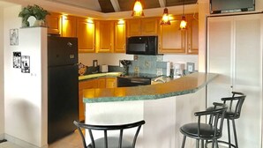 A kitchen with wooden cabinets, a black microwave, a stove, and a bar area with chairs. - #56 Colony One at Sea Mt. Punalu`u (Pahala)