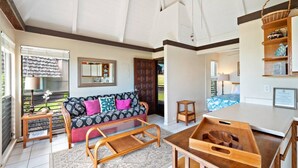 A spacious living area with a sofa, coffee table, and a dining table. There is a small wooden stool and a basket on a shelf. - #04 Rest & Relax at Punalu`u Black Sand Beach (Pahala)