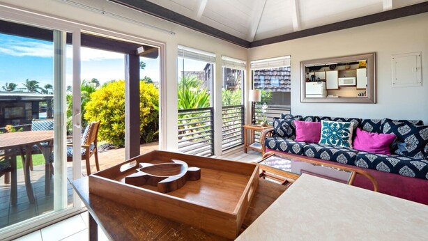 A spacious living area with a wooden coffee table, a sofa with patterned cushions, and a view of greenery through large sliding glass doors. - #04 Rest & Relax at Punalu`u Black Sand Beach (Pahala)