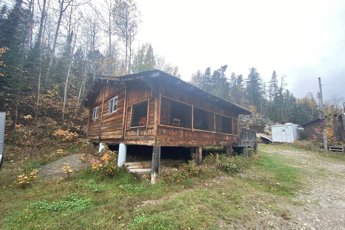 A wooden cabin with a porch, surrounded by trees and a gravel path.