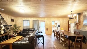 A cozy living room with a leather sofa, wooden coffee table, and a dining area with a table and chairs. A stone fireplace and a kitchen with a stove are visible in the background. - Lake Onaping Lodge (Unorganized North Sudbury)