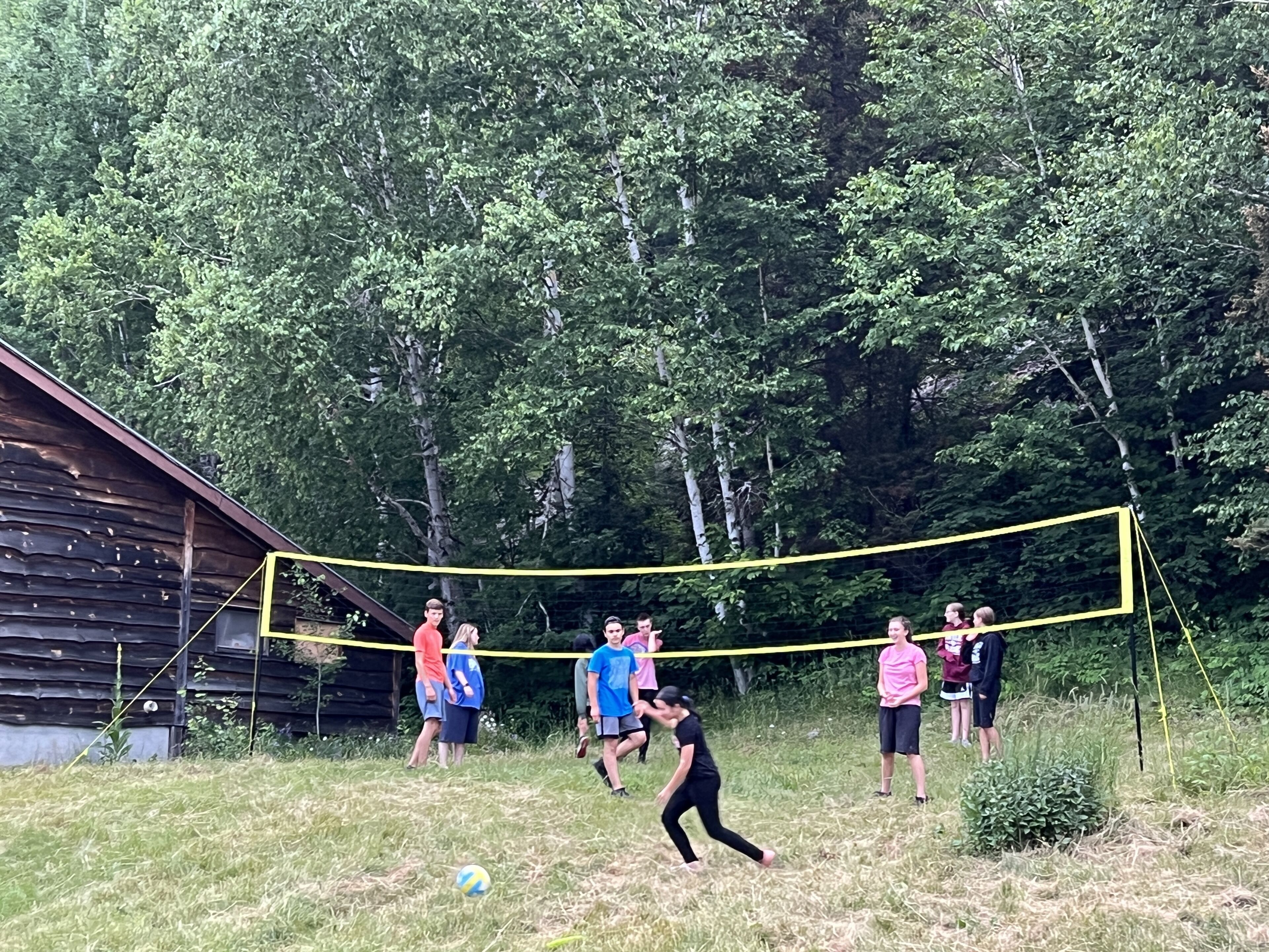 A group of people playing volleyball in a grassy area with a wooden cabin in the background.