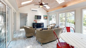 A spacious room with a high ceiling, large windows, and a ceiling fan. There is a dining table with chairs, wicker furniture, and a cabinet with decorative items. - Seabrook Exclusives | 2717 Seabrook Island Rd (Seabrook Island)