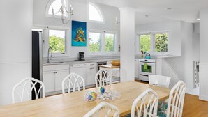 A modern kitchen with white cabinetry, a wooden dining table, and white chairs. There is a painting on the wall, a chandelier, and a window with a view of greenery. - Seabrook Exclusives | 2248 Rolling Dunes (Seabrook Island)