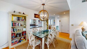 Un amplio espacio habitable con una mesa de comedor de vidrio rodeada de sillas blancas de mimbre, una estantería llena de libros y juegos de mesa, y una lámpara de araña moderna.