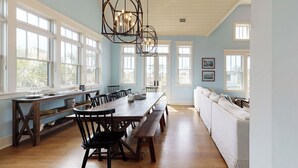Un vaste espace dédié à la salle à manger, avec une longue table en bois et des bancs, entouré de grandes fenêtres et agrémenté d’un canapé blanc.
