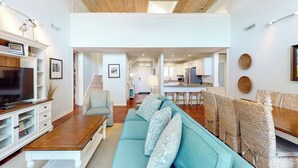 Amplia sala de estar con sofá, televisor y mesa de centro de madera. Una escalera conduce a un nivel superior con cocina y comedor.