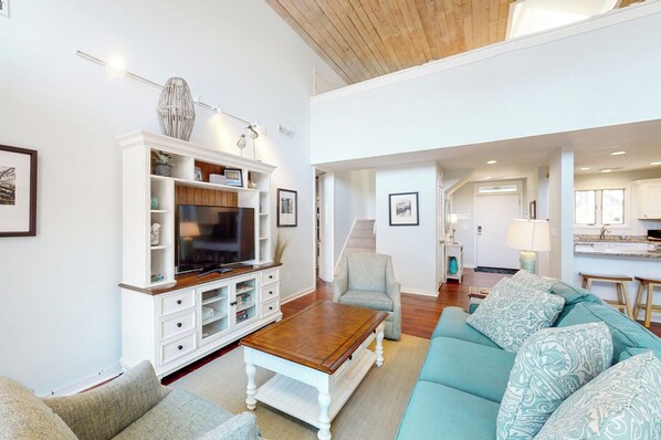 A spacious living room with a large TV, white entertainment unit, teal sofa, and a wooden coffee table. - Seabrook Exclusives | 3637 Pompano Court (Seabrook Island)