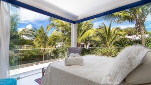 A bedroom with a large window, a bed with white linens, a chair, and a view of palm trees and greenery. - Casa da Árvore - com piscina privativa em Pipa por Carpediem (Tibau do Sul)