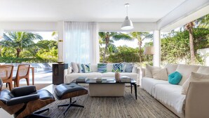 A modern living room with a sofa, coffee table, and chairs. There is a large window with a view of palm trees and greenery. - Casa da Árvore - com piscina privativa em Pipa por Carpediem (Tibau do Sul)