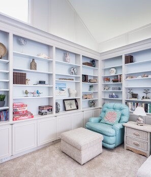 Um quarto bem organizado com estantes brancas embutidas, repletas de livros, objetos decorativos e uma poltrona azul aconchegante.