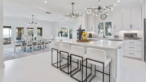 A modern kitchen with white cabinetry, a central island with bar stools, pendant lighting, and a dining area with white chairs and a rug.