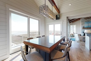 Un comedor amplio con una mesa grande y sillas, ventanas panorámicas con vista a la playa y una lámpara colgante de diseño moderno.