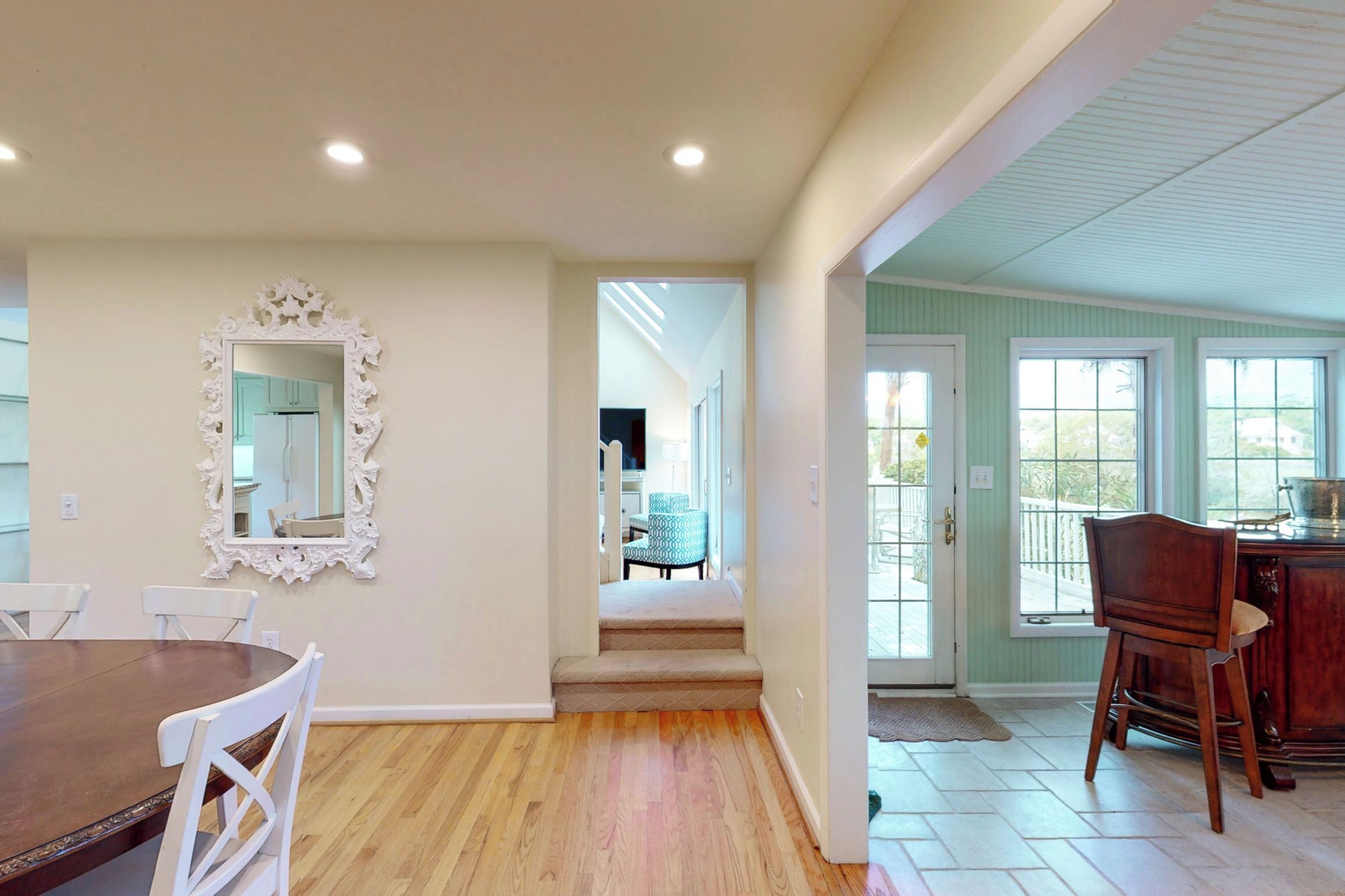 Un comedor con una mesa de madera y sillas blancas, un espejo con un marco decorativo y una escalera que lleva a otra habitación.