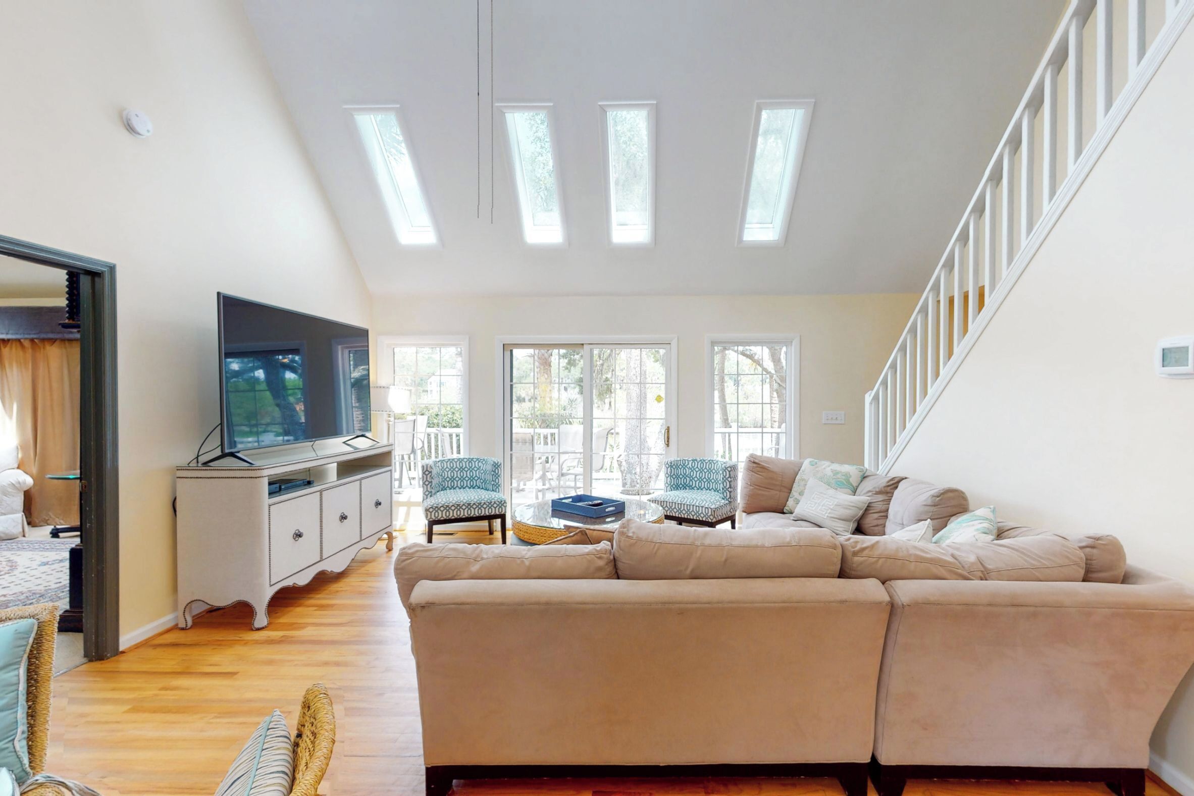 Un salón amplio con un sofá beige grande, dos sillones estampados, una mesa de centro de cristal y un televisor de pantalla plana sobre un mueble blanco. Hay tragaluces en el techo y una escalera a la derecha.