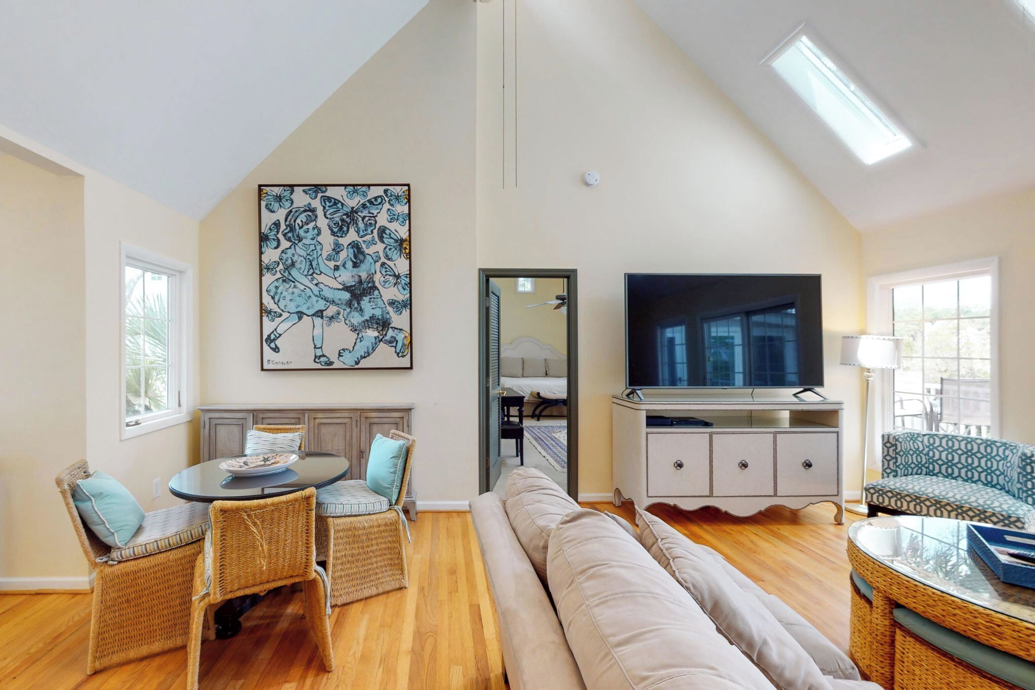 A spacious living room with a high ceiling, a large flat-screen TV, wicker furniture, and a painting on the wall.