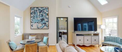 A spacious living room with a high ceiling, a large flat-screen TV, wicker furniture, and a painting on the wall.