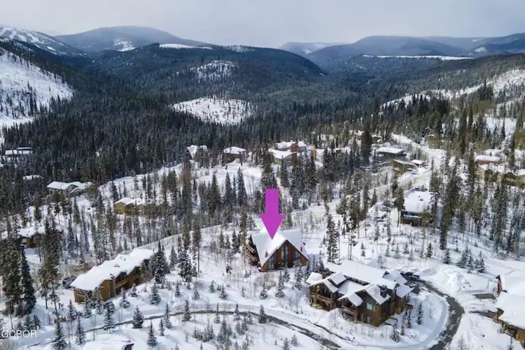 Un chalet enneigé, doté de plusieurs cabanes, entouré d’une forêt dense et de montagnes.