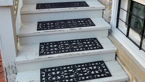 Un conjunto de escalones blancos de madera con rejas metálicas negras decorativas, que conducen a una barandilla blanca y un muro de ladrillo.