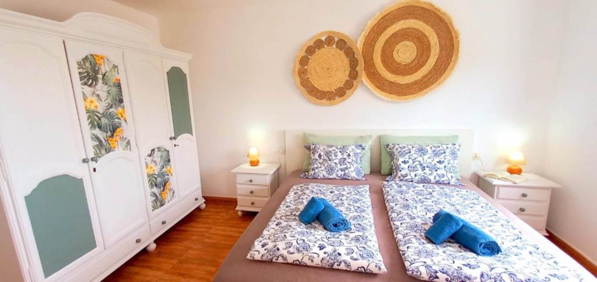 Un lit double avec une literie à motifs floraux, deux tables de chevet avec des lampes, une armoire avec des panneaux à motifs floraux et deux éléments de décoration murale.