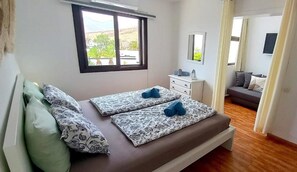 Ein Schlafzimmer mit Bett, Fenster, Nachttisch, Kommode und Blick ins Freie.