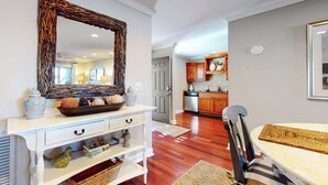 Una habitación bien iluminada con una mesa de comedor de madera, una consola blanca con objetos decorativos, y una cocina visible a través de una puerta abierta.