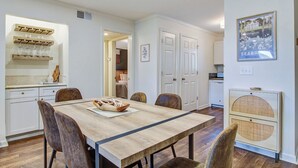A dining area with a wooden table and chairs, a kitchen with white cabinets, and a framed picture on the wall. - Seabrook Exclusives | 2011 Marsh Walk (Seabrook Island)