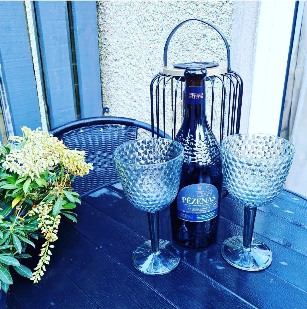 A bottle of Pêzenas wine, two crystal glasses, and a wicker chair on a wooden table.