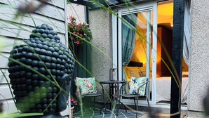 Un rincón exterior acogedor con una mesa y sillas, una escultura decorativa, y vistas a una habitación cálidamente iluminada a través de un ventanal.