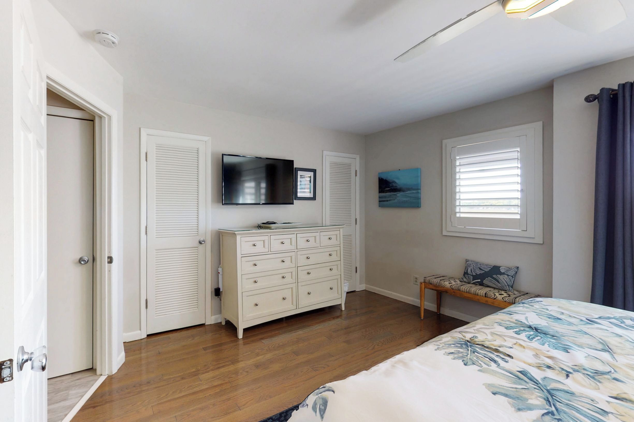 Une chambre à coucher avec un lit, une commode, un téléviseur et une fenêtre avec des stores.