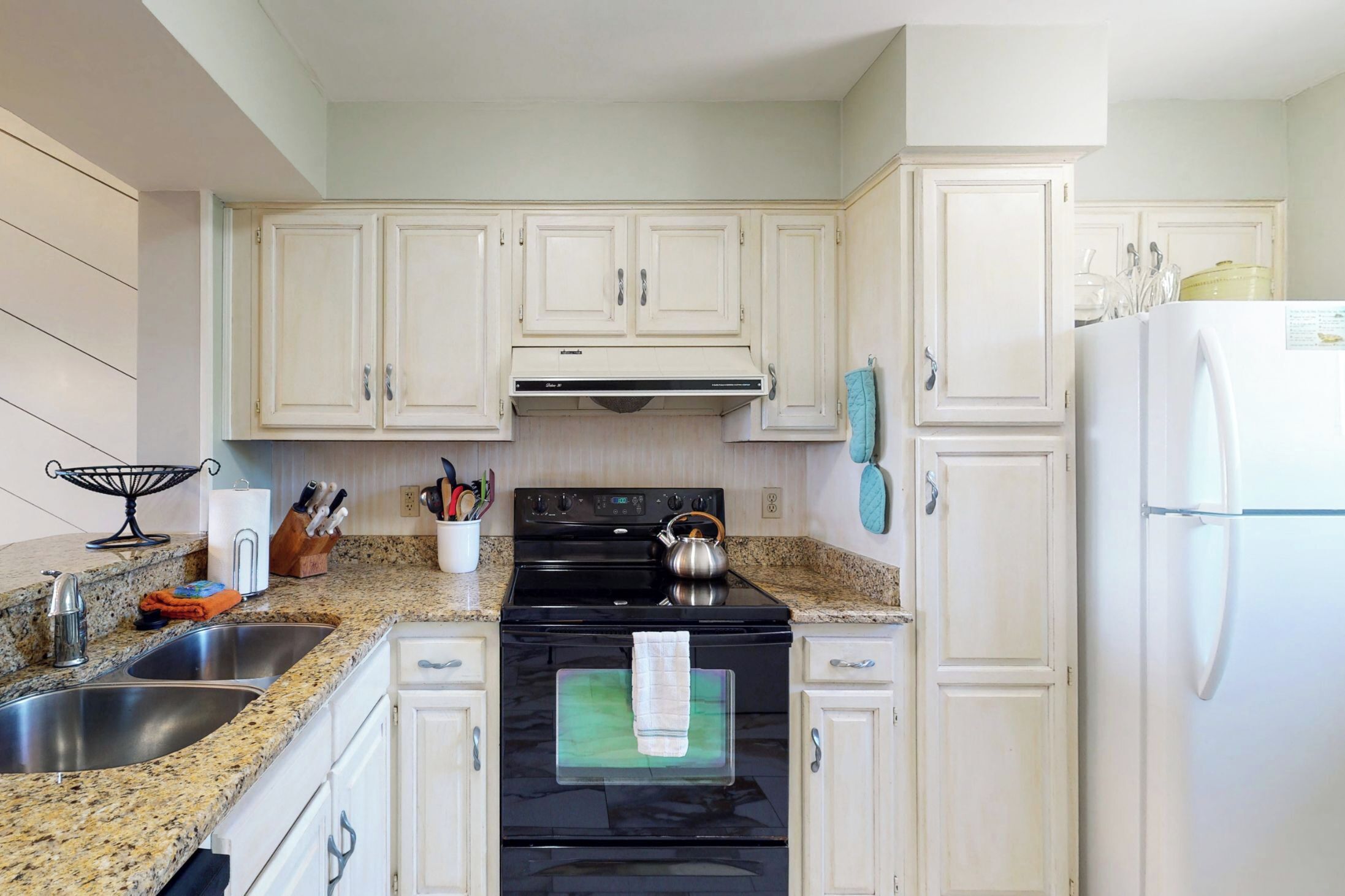 A kitchen with white cabinets, a black stove, stainless steel sinks, and a white refrigerator.