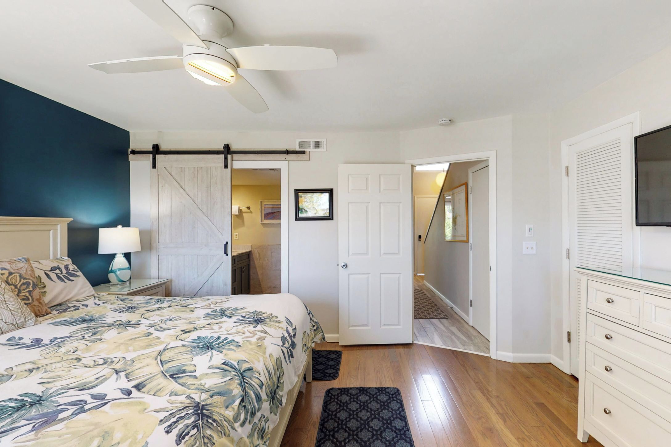 Un dormitorio con una cama, un ventilador de techo, una mesita de noche con lámpara, una puerta y un pasillo que conduce a otra habitación.