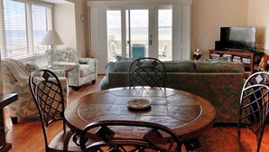 A living room with a round wooden table, two chairs, a sofa, a television, and a balcony with a view. - Seabrook Exclusives | 1302 Pelican Watch (Seabrook Island)