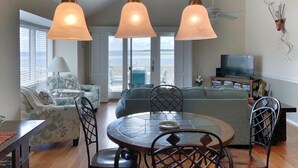 A spacious living area with a dining table, chairs, and a sofa. There is a television and a bookshelf in the background. - Seabrook Exclusives | 1302 Pelican Watch (Seabrook Island)