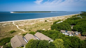 Vue aérienne d’une zone côtière avec une plage, un chemin de terre et plusieurs bâtiments entourés de verdure.