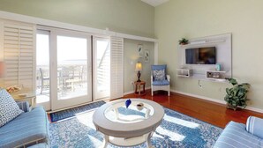 Un salon doté d’une grande porte-fenêtre coulissante donnant sur un balcon, d’une table basse ronde, d’un téléviseur fixé au mur et d’un tapis bleu à motifs.